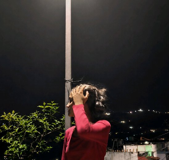 Profile view of a woman in a red top resting her hand on her head beside a streetlamp, overlooking a lit city at night.