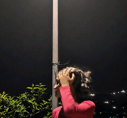 Side profile of woman in pink top touching her hair outdoors on a dark night, distant city lights visible.