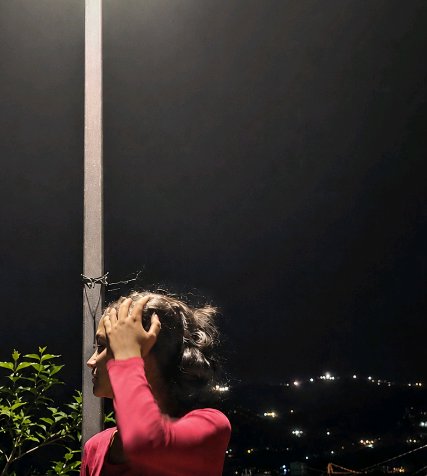 Side profile of a woman in a pink top adjusting her hair under a streetlight overlooking a lit hi...