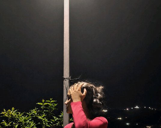 Side profile of a woman in a pink top resting her hand in her hair beside a street lamp overlooki...