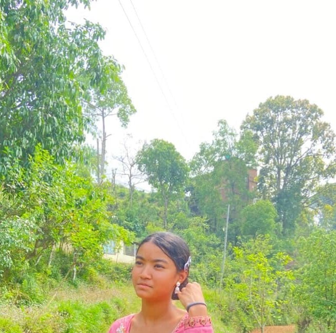 Young woman in a pink top poses outdoors against a lush, tree-filled rural hilly village landscape