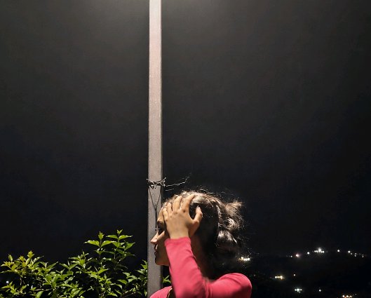Side profile of a woman in a pink top adjusting her hair next to a streetlight, dark night sky and distant city lights in ...