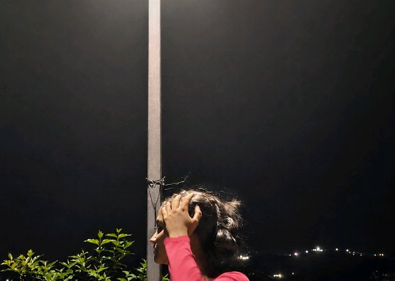 [Side profile of woman in pink top adjusting hair beside a street lamp post on a dark night, distant town lights in the ba...