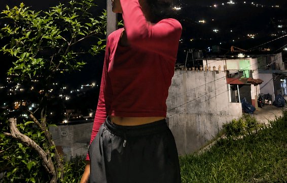 [Partially framed woman in a red cropped top standing on a grassy hillside overlook at night, with a lit residential neigh...