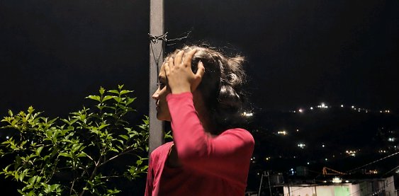 Profile view of a woman in a red top touching her hair, overlooking a lit hillside from a balcony...