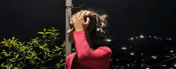 Profile of a woman in a pink top touching her wind-tousled hair, overlooking a twinkling hilly ci...