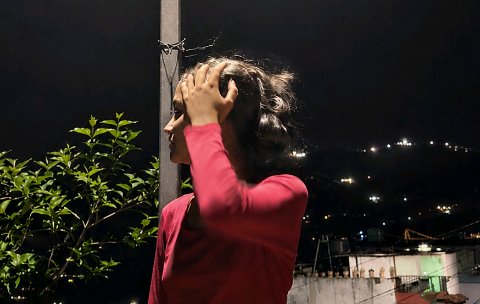 Side profile of a woman in a pink top touching her hair, standing on a balcony overlooking a lit ...