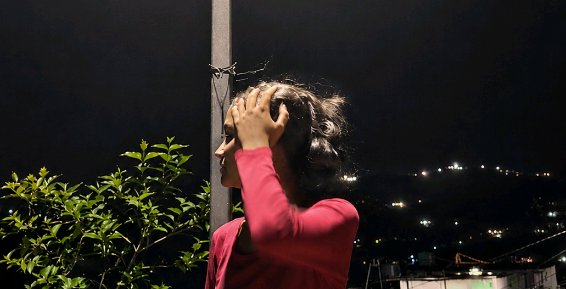 Side profile of a woman in a pink top touching her hair, overlooking a lit hillside city from a b...
