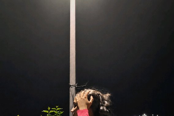 [Profile of a young woman touching her hair under a glowing streetlight against a dark night sky, small green foliage in t...