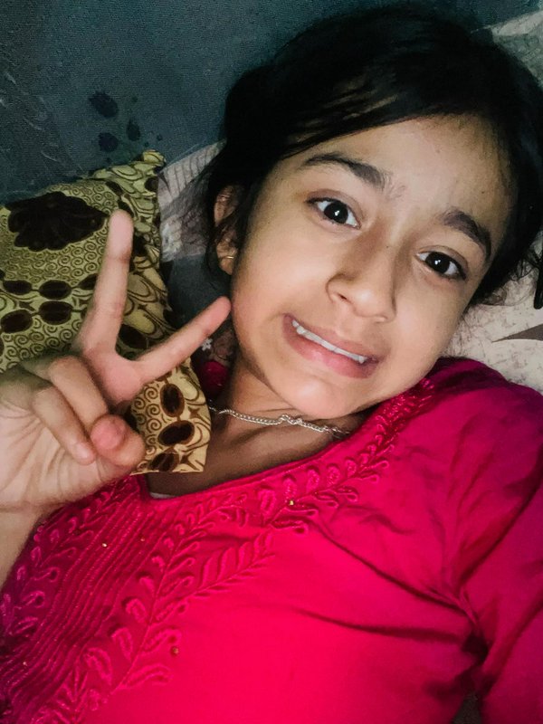 Smiling young girl in a bright embroidered red top takes a cozy bedroom selfie holding up a peace...