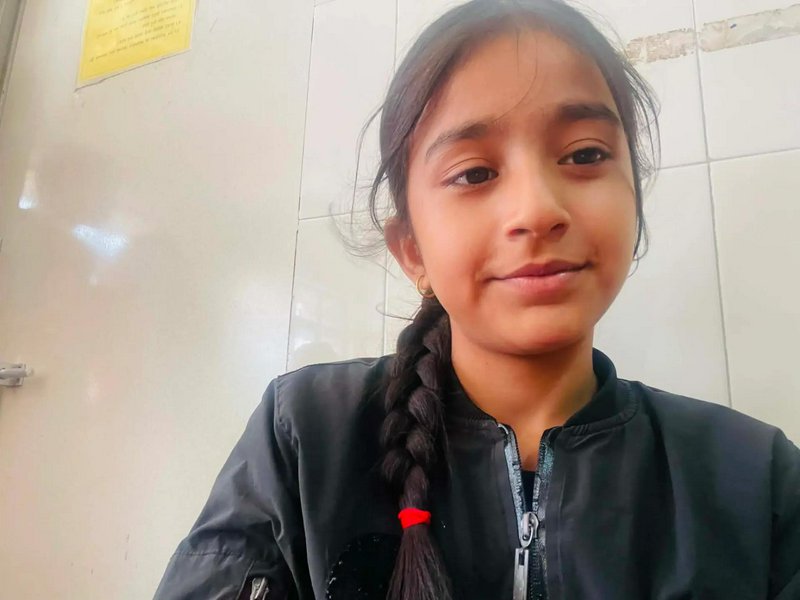 Close-up indoor selfie of a young girl with a long braid, soft smile, and black zip-up jacket