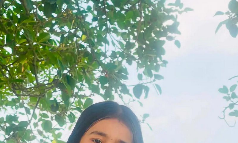 Partial view of young woman with bindi peeking into frame, below lush green tree foliage against ...