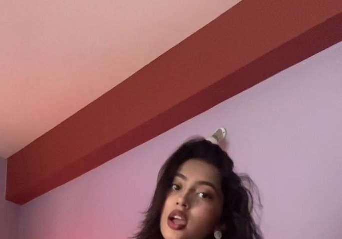 Partial low-angle selfie of a young woman with dark wavy hair, playful expression, against pastel...