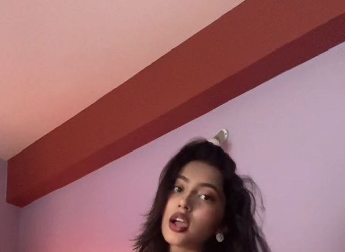 Low angle candid selfie of a young woman with tousled dark hair making a playful face, against pa...