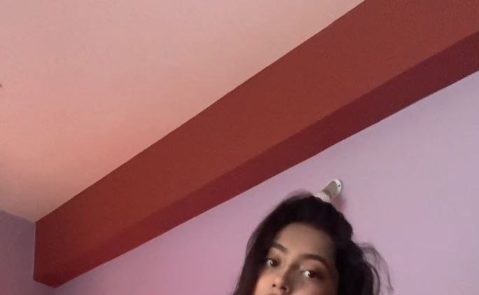 Low-angle candid selfie of a young woman with dark wavy hair, with pastel pink ceiling and red be...