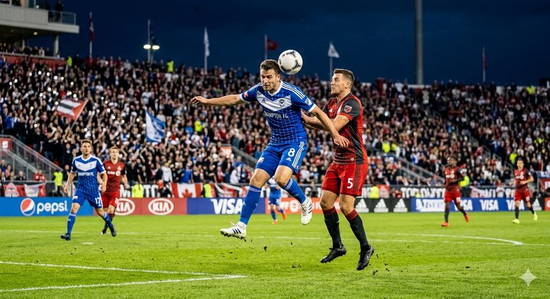 Two soccer players in blue and red jump for a header during a night match in a packed stadium.