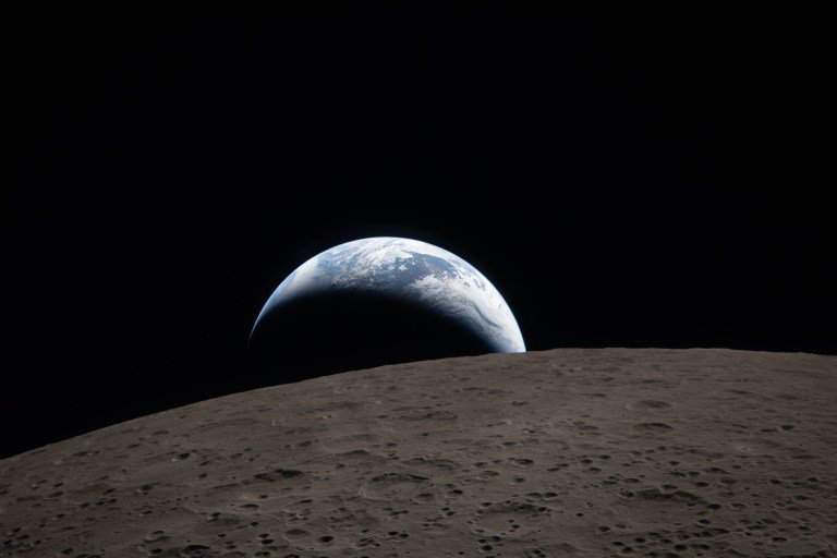Glowing crescent Earth rises above the heavily cratered grey lunar horizon against pitch black de...