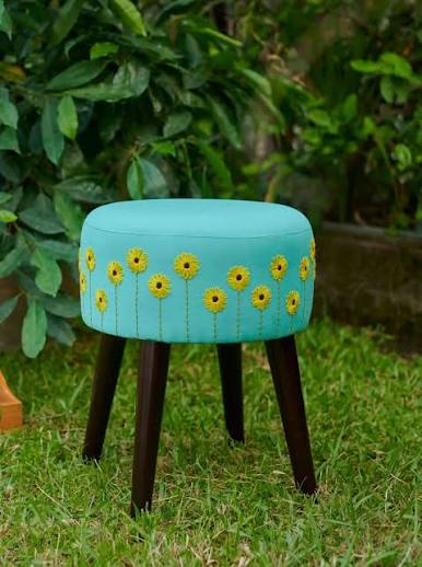 Turquoise upholstered stool with yellow sunflower embroidery in a garden setting.