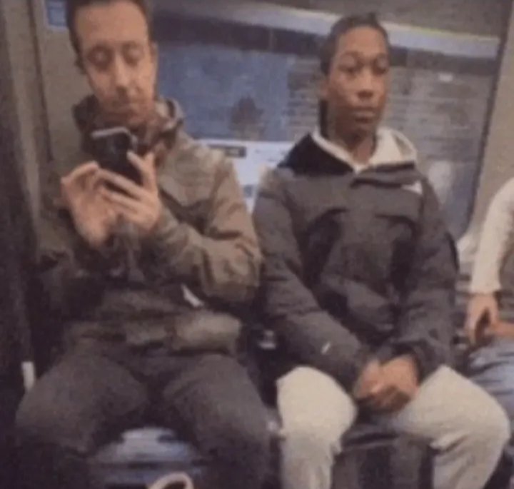 Two men sit on a subway train; one is taking a selfie with his phone while the other looks ahead.
