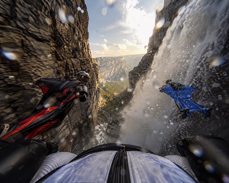 First-person POV of wingsuit flyers gliding through a narrow rocky mountain canyon beside a casca...