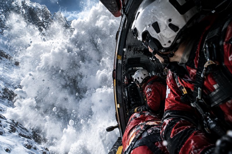 Mountain rescue team inside an open helicopter observes a massive cascading avalanche on alpine peaks