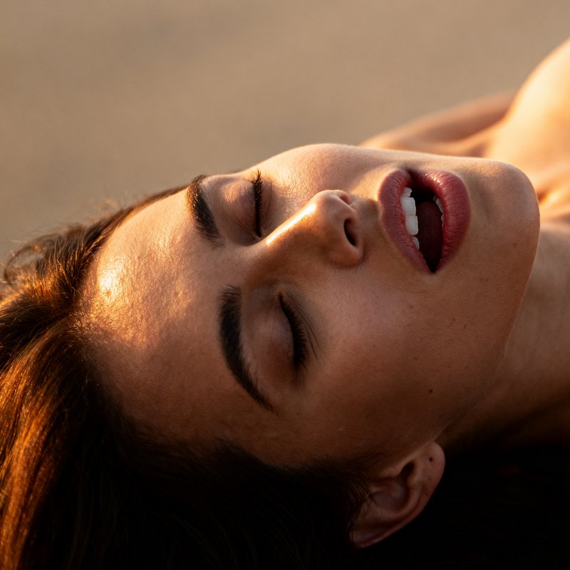 Close-up of a woman with eyes closed, sunlit face and relaxed expression on a beach.
