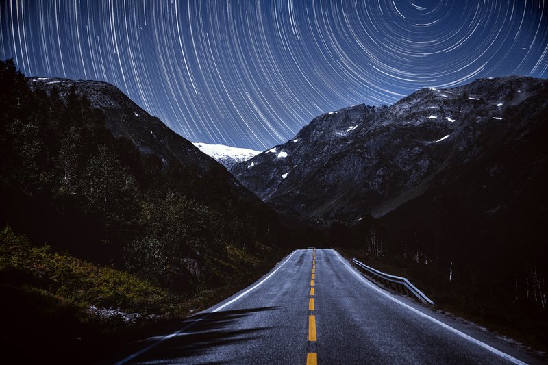 Nighttime mountain road under star-trail sky in long-exposure night photography.