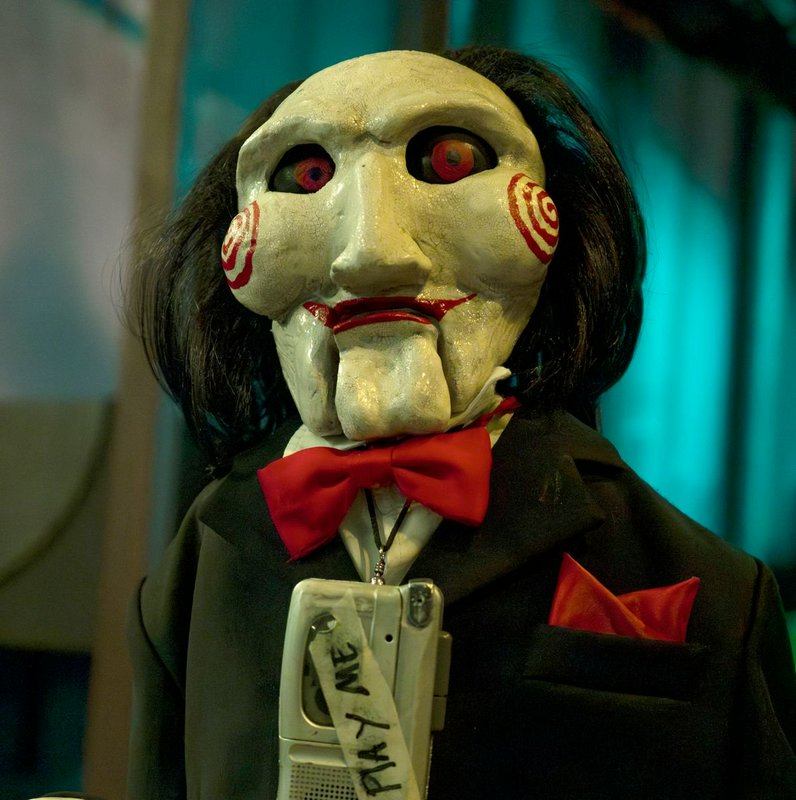 Creepy ventriloquist dummy with a white mask, red spiral cheeks, black suit, and red bow tie.