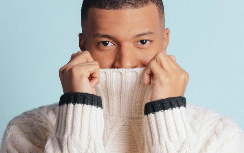 Person in a cream cable-knit sweater pulls the collar up against a light blue background.