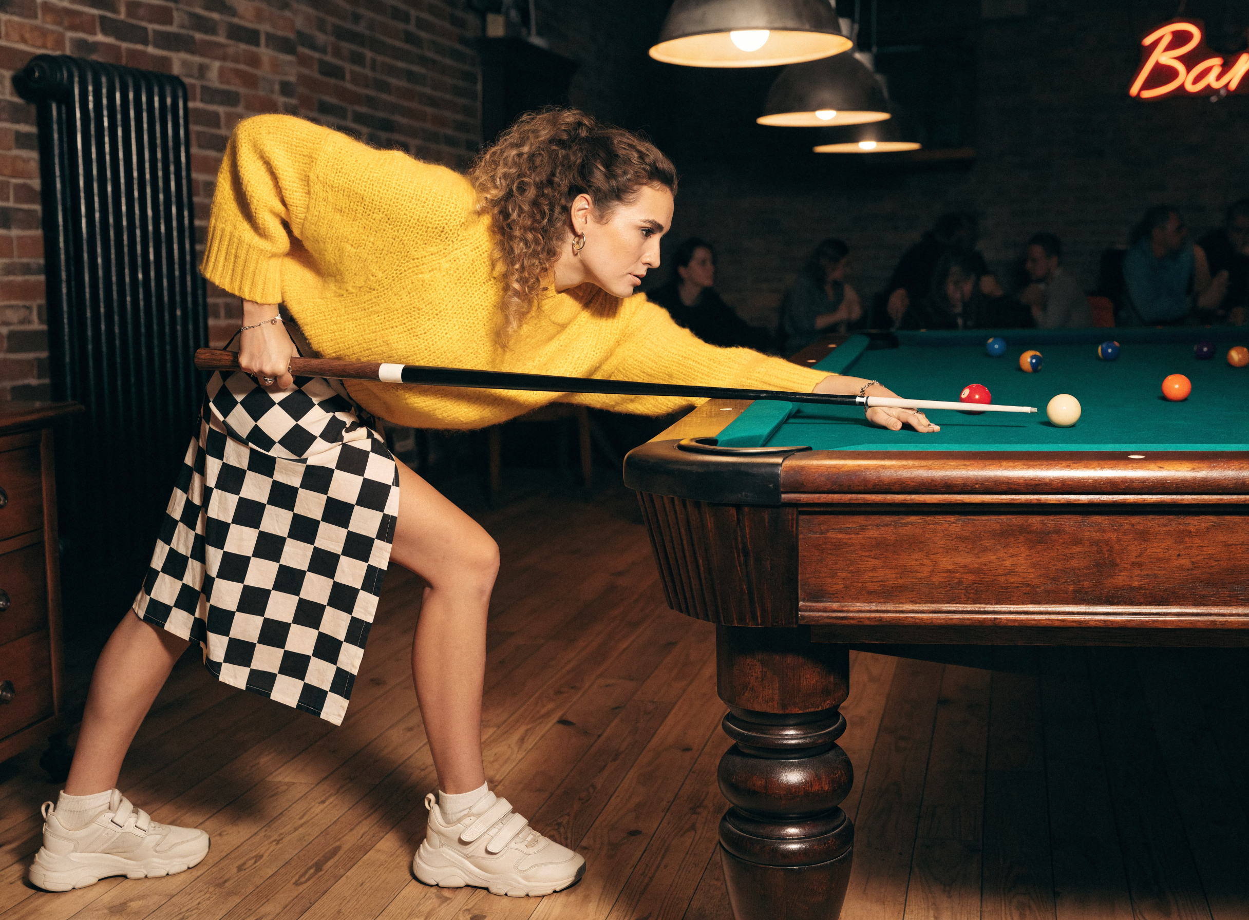 Focused woman in a yellow knit sweater lines up a pool shot over a green felt billiards table in a cozy brick bar.