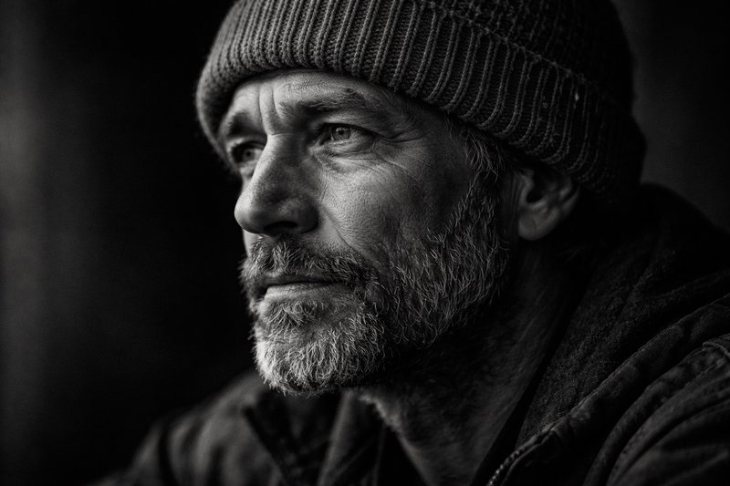 A black-and-white portrait of a bearded man wearing a knit beanie, looking pensively to the left.