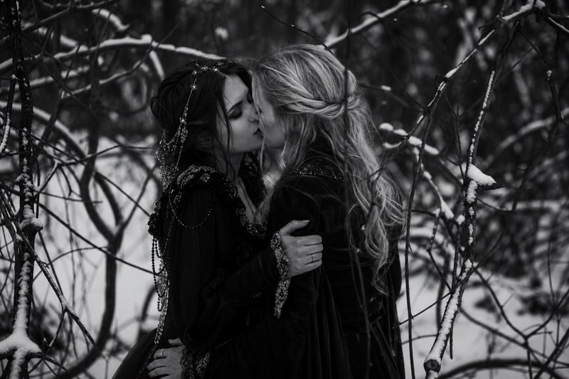 Black and white photo of two women in ornate gothic gowns sharing a kiss in a snow-dusted winter forest.
