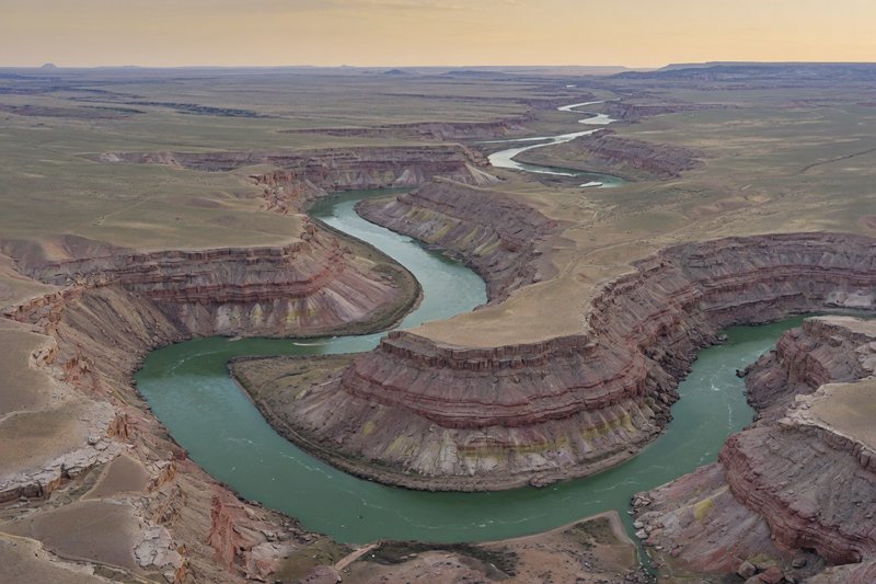 [Aerial view of a winding green river carving tight meanders through layered red sandstone canyon badlands at hazy golden ...