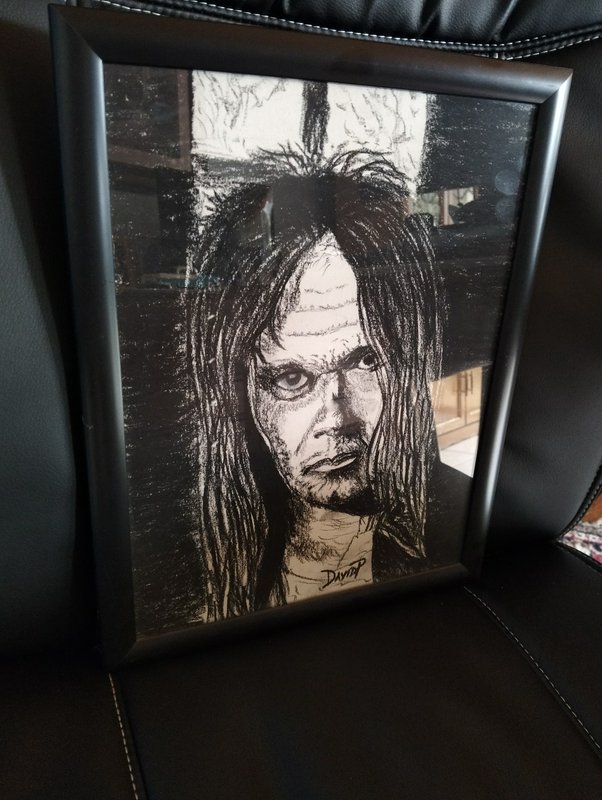Framed black and white charcoal portrait of a gaunt long-haired eerie figure propped on a black leather chair