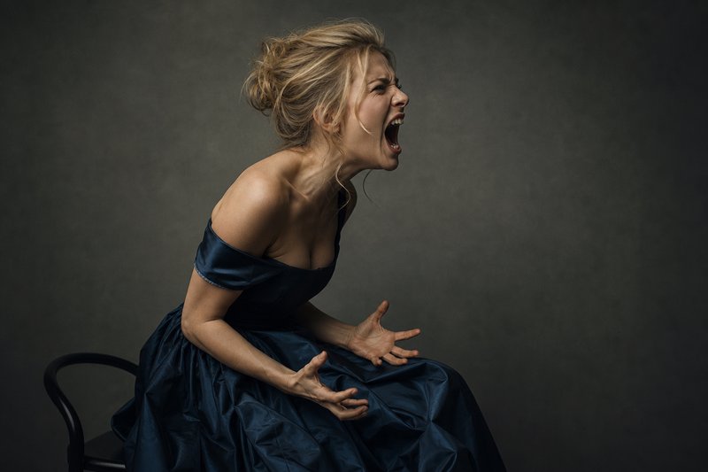 Frustrated blonde woman in a dark satin gown shouting while seated for a moody studio portrait