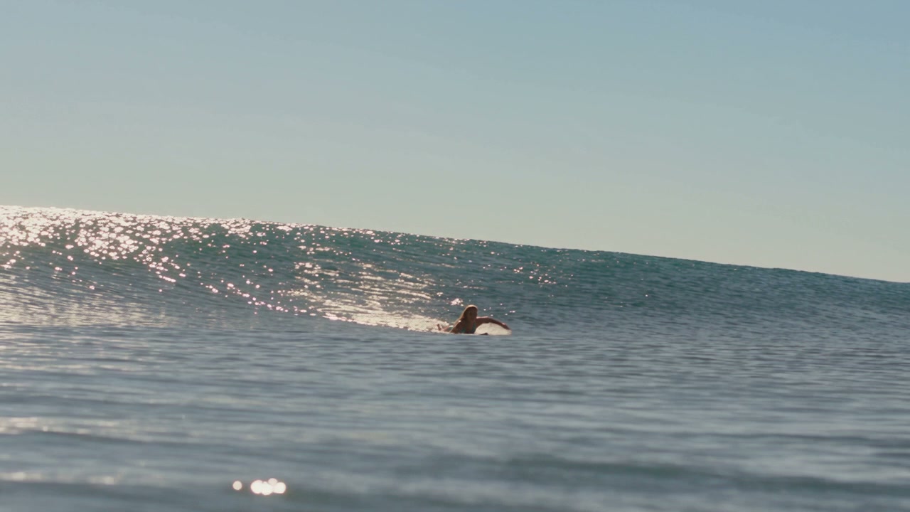 Surfer paddles on a surfboard beside a sunlit glistening ocean wave under a clear pale blue sky.