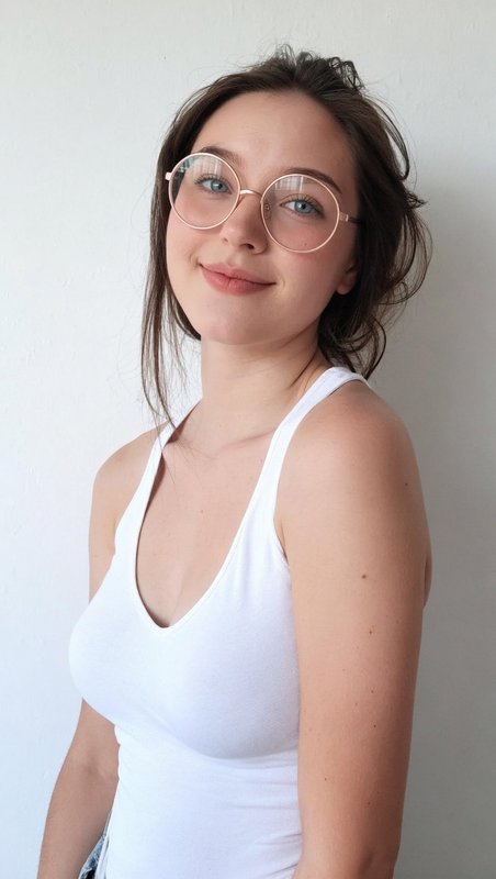 Young woman wearing round glasses and a white tank top, posing softly against a light wall.
