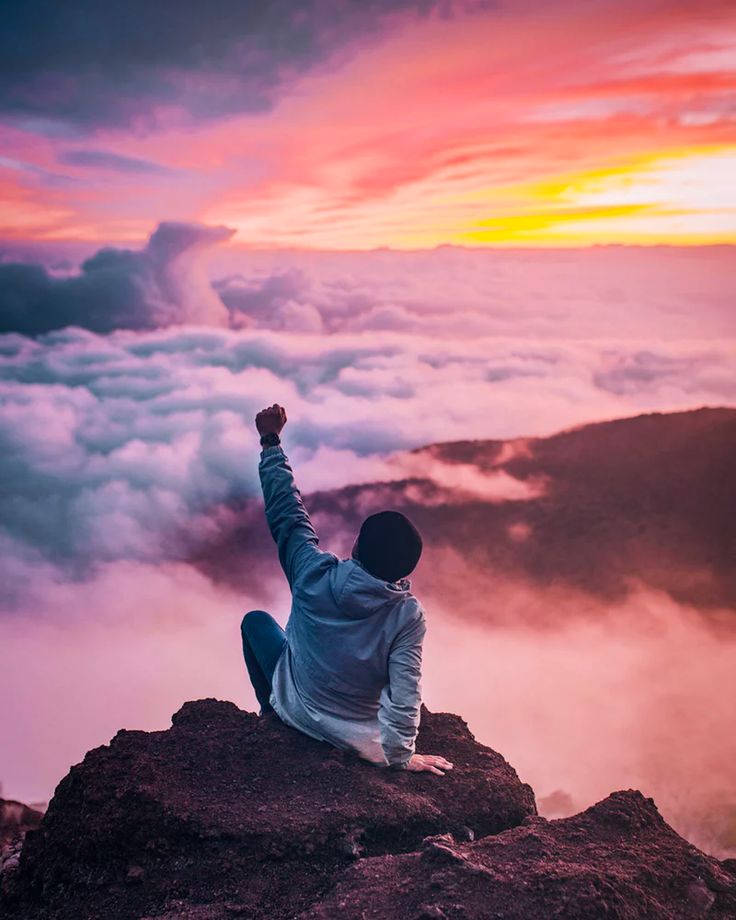 Person sitting on a rocky cliff above clouds at sunset, fist raised in triumph.