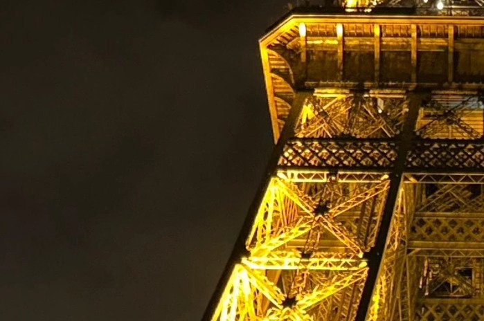 Low-angle close-up of the golden illuminated Eiffel Tower, showcasing its lattice ironwork against a dark night sky.