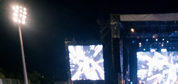 Blurry nighttime outdoor concert view featuring glowing LED stage screens and a bright stadium fl...
