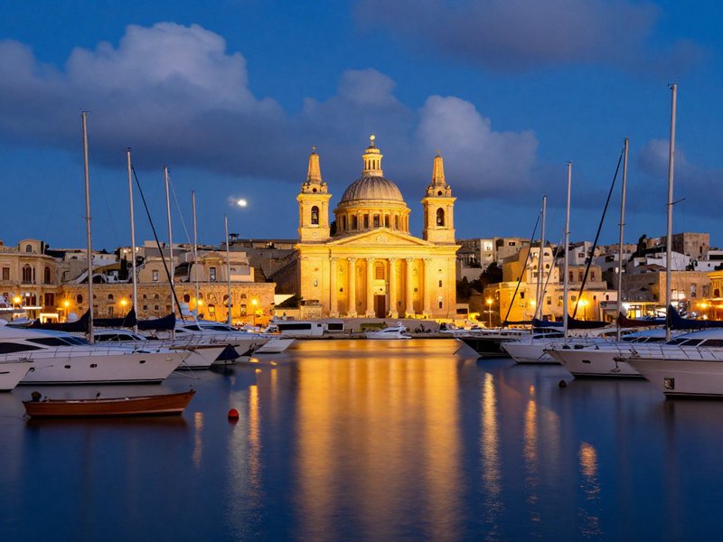 Night view of a lit baroque cathedral on the waterfront with boats in a calm harbor.