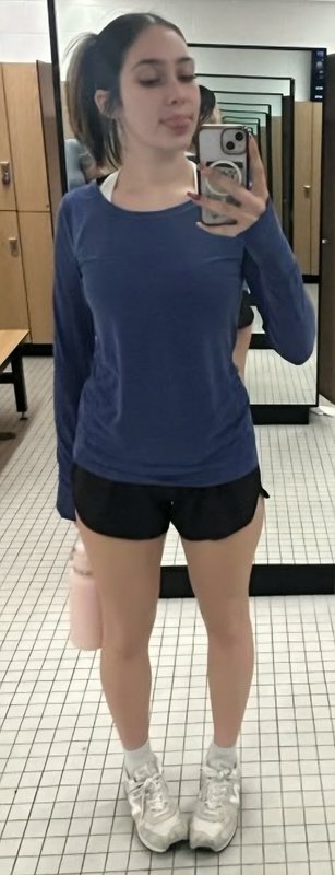 Young woman in a blue long-sleeve athletic top and black shorts taking a mirror selfie in a gym l...