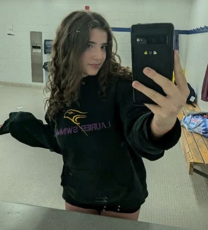 Young woman taking a mirror selfie in a locker room, wearing a black hoodie with a colorful sport...