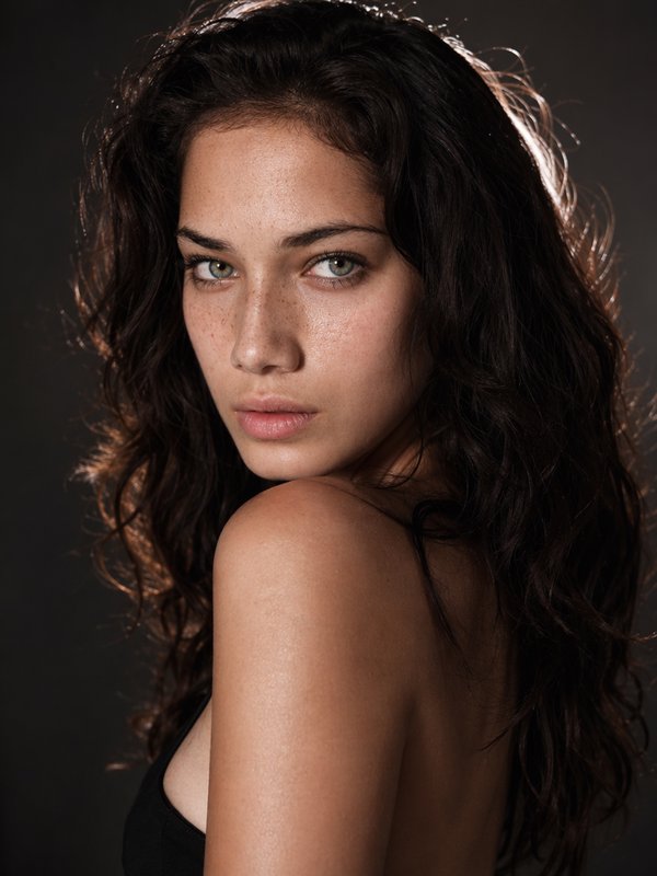 Portrait of a young woman with dark wavy hair and freckles, looking over her shoulder against a dark backdrop.