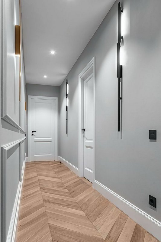 Modern grey hallway with white paneled doors, chevron wood floor, and vertical wall sconces.