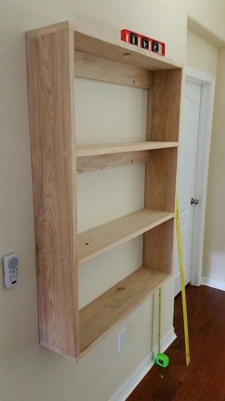 Unfinished recessed wooden wall shelf mid-installation, with a level tool and tape measure used f...