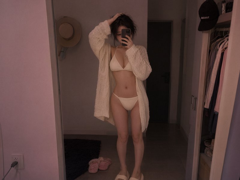 Young woman takes casual mirror selfie wearing cream bikini and oversized knit cardigan in dim co...