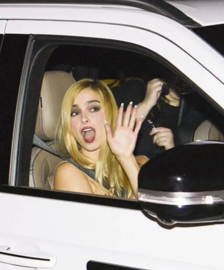 Blonde woman with a playful surprised expression waves from the passenger seat of a white SUV at night.