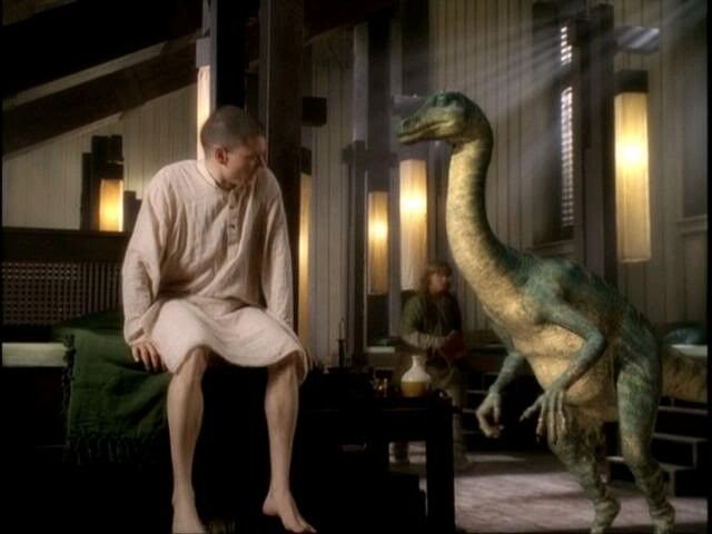 A man in a pale linen nightgown sits on a bed face-to-face with a small long-necked green dinosau...