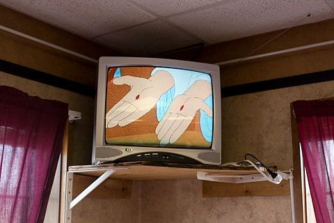 Vintage CRT tube TV mounted to a high wall shelf displays a 2D cartoon of outstretched hands, fla...
